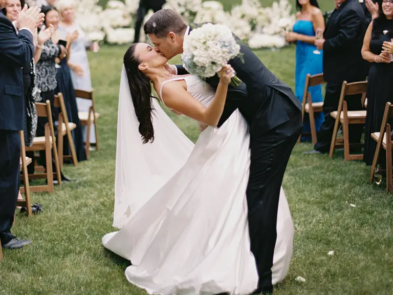 A bride and groom kiss in the aisle as guests applaud on the club lawn